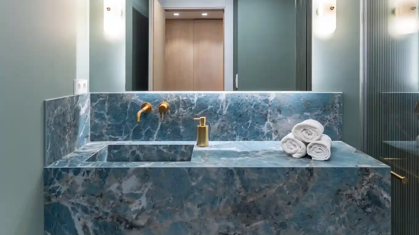 Baño con encimera y frontal de mármol azul, grifería dorada y espejo retroiluminado, uno de los ambientes del catálogo de la web de Ascale
